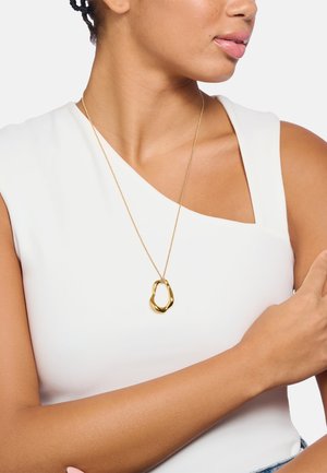 Goldanhänger-Halskette mit einer organischen, abstrakten Form an einer zarten Kette, getragen über einem weißen Oberteil, mit glatter Textur und Glanz.