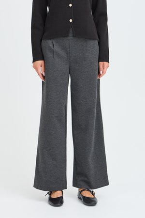 Femme portant un pantalon large gris foncé, des ballerines noires et un top noir boutonné, debout devant un fond uni.
