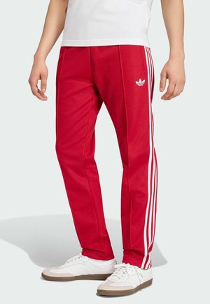 Muž v červených teplácích Adidas s bílými pruhy a logem, doplněný bílými teniskami a bílým tričkem.