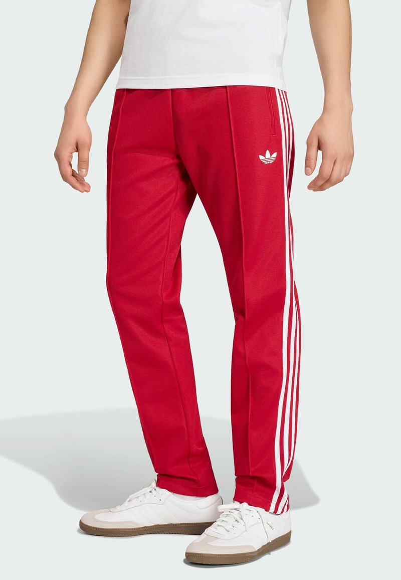 Homme portant un pantalon de survêtement rouge Adidas avec des bandes blanches et un logo, associé à des baskets blanches et une chemise blanche.