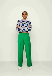 Suéter curto multicolorido com padrões geométricos em azul, roxo e verde, combinado com calças verdes de cintura alta e ténis brancos.