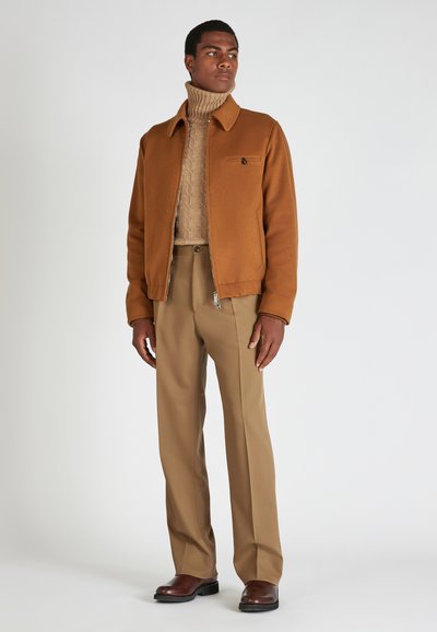 Braune, Reißverschluss-Jacke über einem strukturierten beigen Rollkragenpullover, kombiniert mit hellbraunen Hosen und dunklen braunen Schuhen. Schlichtes Design ohne Verzierungen.