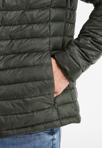 Chaqueta acolchada verde con patrón de acolchado horizontal, que presenta un bolsillo lateral y una textura suave y brillante.