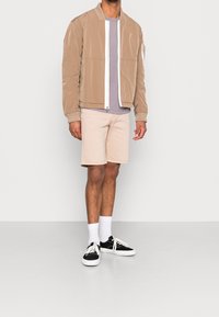 Beige bomber jacket with white zipper, layered over a light purple shirt. Paired with beige shorts and black sneakers with white laces.