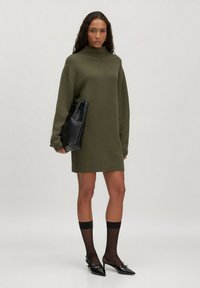 Olivengrünes gestricktes Kleid mit hohem Kragen und langen Ärmeln; kombiniert mit schwarzen spitzen High Heels und einer schwarzen Handtasche. Durchsichtige schwarze knielange Socken.