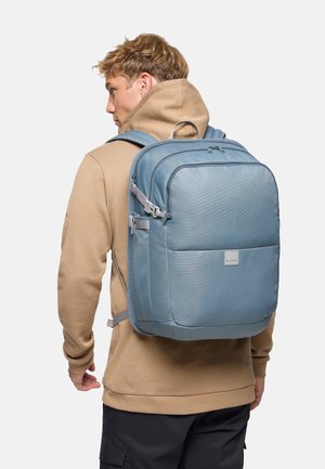 Mann in beigefarbenem Hoodie mit großem blauem Rucksack mit mehreren Fächern, stehend vor hellem, einfarbigem Hintergrund.