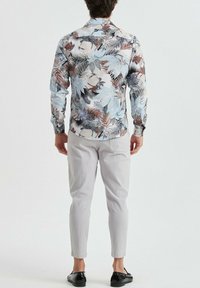 Chemise à manches longues florale bleu clair, ornée de divers motifs de feuilles en brun foncé et vert, associée à un pantalon effilé gris clair et des chaussures noires.