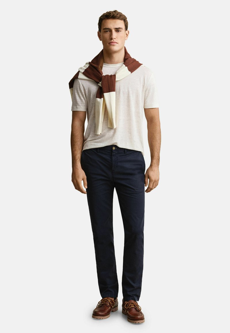 Joven con camiseta beige clara, pantalones azul marino oscuro, suéter marrón con rayas blancas sobre los hombros y zapatos de cuero marrón.