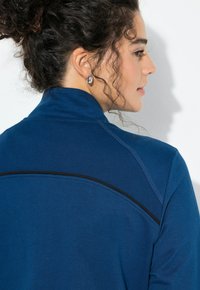 Chaqueta deportiva azul con un cuello alto, tela texturizada y una franja negra decorativa en la espalda. Acabado liso y diseño ajustado.