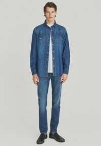 Giovane uomo in piedi, con una camicia di jeans blu aperta sopra una maglietta bianca, jeans blu e scarpe nere, su uno sfondo chiaro e neutro.