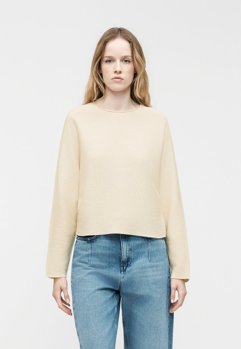 Jeune femme aux longs cheveux blonds portant un haut crème à manches longues uni et un jean bleu taille haute, debout devant un fond blanc.