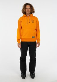 Mann mit lockigem Haar, der einen orangefarbenen Hoodie, schwarze Hosen und schwarze Skischuhe trägt, steht vor einem weißen Hintergrund.