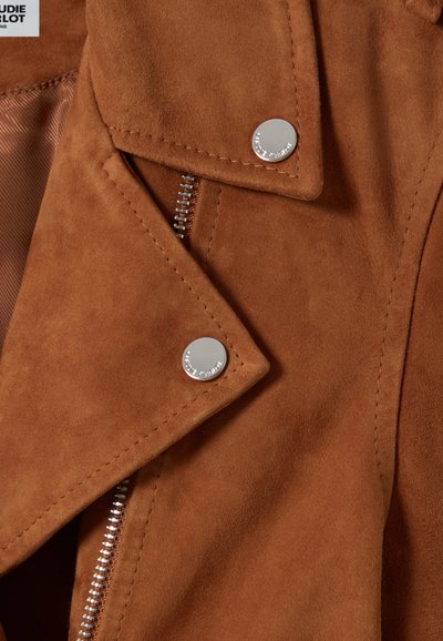 Giacca in suede marrone con dettagli in metallo argentato, dotata di una zip diagonale e bottoni a scatto; primo piano che mette in risalto la texture e i dettagli di cucitura.