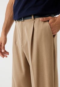 Tan plissébroek van gladde stof, met zijzakken en een donkere riem met een vierkante metalen gesp. Zichtbare hand in de zak.