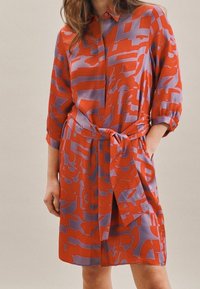 Mujer que lleva un vestido de manga larga hasta la rodilla con patrones abstractos en naranja y púrpura y un cinturón a juego, con una mano en el bolsillo.
