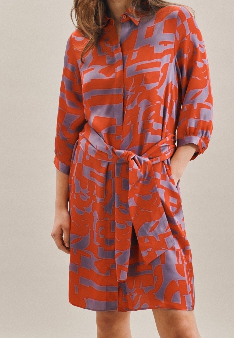 Mujer que lleva un vestido de manga larga hasta la rodilla con patrones abstractos en naranja y púrpura y un cinturón a juego, con una mano en el bolsillo.