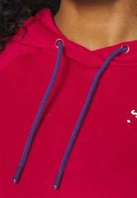 Červená mikina s kapucí s fialovými šňůrkami, měkkou texturou látky a malým bílým logem. Výstřih je ležérní s volným střihem.