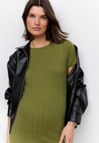 Olivengroene geribbelde jurk met korte mouwen, gecombineerd met een glanzend zwarte leren jas met een zilveren ritssluiting en textuurstof.