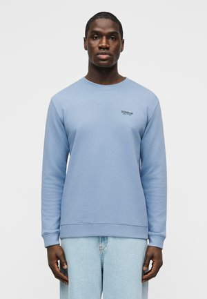 Hombre con sudadera de cuello redondo azul claro con el logo "DONDUP Made in Italy" en el pecho y jeans azul claro, sobre un fondo sencillo.