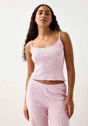 Kvinde iført lys pink ribstrikket camisole og matchende bukser med små røde hjerteprints mod en ensfarvet lys baggrund.