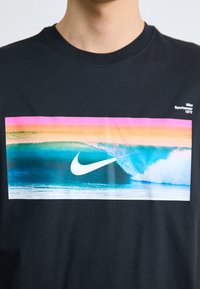 Svart bomull T-skjorte med et stort rektangulært grafikk av havbølger i teal, rosa og oransje, med en hvit Nike-logo nederst.