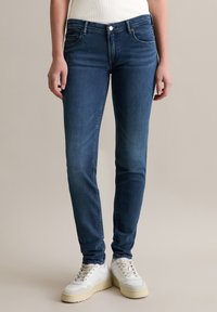 Jean en denim extensible bleu foncé avec poches avant, fermeture éclair et léger délavage aux cuisses. Associé à des baskets blanches montantes.