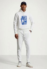 Άνδρας που φοράει λευκή πόλο φούτερ με μπλε λογότυπο, ταιριασμένα λευκά παντελόνια, λευκά αθλητικά παπούτσια, ναυτικό καπέλο και ρολόι, στέκεται μπροστά από απλό φόντο.