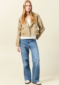 Beige cropped double-breasted jacket met grote knopen, gedragen over een witte trui en gecombineerd met brede blauwe jeans. Glad textuur van de stof.
