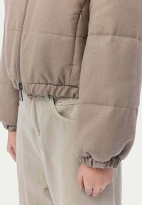 Brun quiltad jacka med dragkedja, elastiska ärmslut, tillsammans med ljusbeige byxor med framfickor och en strukturerad yta.