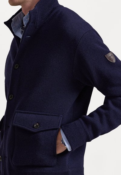 Μπλε πλεκτή ζακέτα με ψηλό γιακά, δύο μπροστινές τσέπες, μαύρα κουμπιά και διακόσμηση με σήμα στο αριστερό μανίκι. Απαλή υφή σε όλη την επιφάνεια.