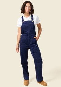 Marineblaue Cord-Overall mit verstellbaren Trägern, Fronttasche und seitlichen Knöpfen, getragen über einem weißen T-Shirt, kombiniert mit beigefarbenen Slippern.