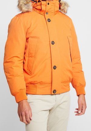 Helles orangefarbenes Winterjacke mit pelzbesetzter Kapuze, Knopf- und Reißverschluss, Seitentaschen und gerippten Bündchen, getragen über hellbeigen Hosen.