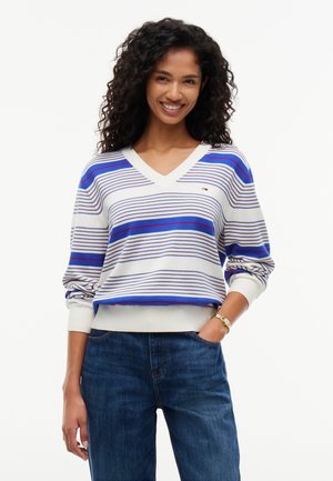 Kvinde med krøllet hår smiler, iført en hvid V-hals trøje med blå og røde striber og blå jeans, den ene hånd i lommen.