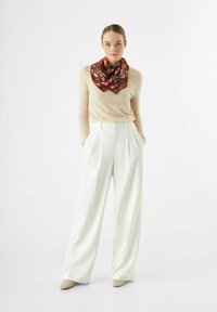 Pull beige, pantalon large blanc et une écharpe en soie rouge à motif nouée au cou. Textures lisses, coupe décontractée.