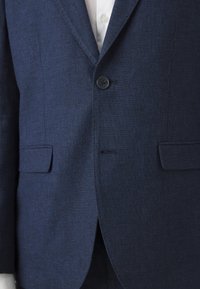 Σκούρο μπλε σακάκι φτιαγμένο από υφασμάτινη ύφανση, με κλείσιμο με μονό κουμπί, δύο μπροστινές τσέπες και σχέδιο με γιακά που σχηματίζει γωνία.