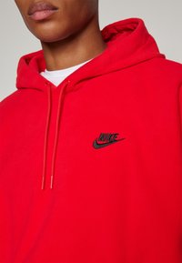Felpa rossa in pile con cappuccio, dotata di cordino regolabile e logo Nike nero sul petto. Il tessuto appare morbido e leggermente testurizzato.