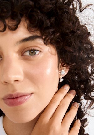 Nainen, jolla on kiharat tummat hiukset ja auringonmuotoiset hopeiset korvakorut, koskettaa hellästi korvaansa, taustalla neutraali väri.