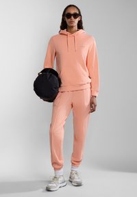 Felpa rosa con cappuccio e tasca frontale, abbinata a pantaloni della tuta rosa con fianchi. Il modello tiene in mano una borsa circolare nera.