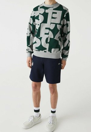 Mann trägt grünes und graues gemustertes Sweatshirt, marineblaue Shorts mit Logos, weiße Sneakers und weiße Socken mit dunklen Streifen, steht vor einfarbigem Hintergrund.