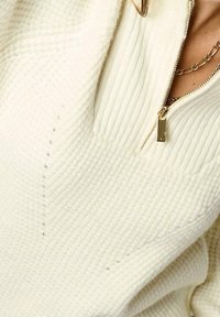 Haut à fermeture éclair couleur crème avec texture côtelée, col haut et petit détail de fermeture éclair dorée. Présente des accents de couture subtils.