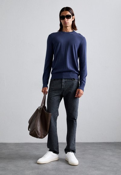 Marineblå strikkegenser, mørke jeans, hvite joggesko og en brun skinnveske. Modellen har på seg svarte solbriller og står mot en nøytral bakgrunn.