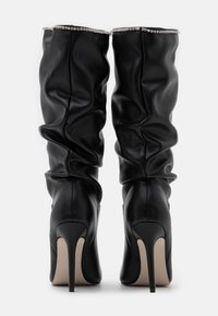 Botas negras holgadas hasta la rodilla con acabado de cuero liso y tacones puntiagudos. Acentuadas con una fila de piedras brillantes en la parte superior.