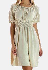 Crèmekleurige jurk met korte, geplooide mouwen, een gerimpelde taille en knoopdetails bij de halslijn; gladde textuur, knielange silhouet.