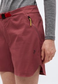 Μπορντό αθλητικό σορτς με κόκκινη ρυθμιζόμενη ζώνη και τσέπη με φερμουάρ, φορεμένο με γκρι μακρυμάνικο τοπ, εμφανίζοντας το δεξί χέρι σε χαλαρή στάση.