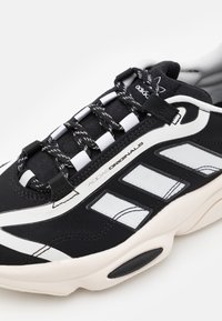 Svart sportskor med vita accenter, övre del i tyg, distinkt tredelad design, strukturerade skosnören och gummiyttersula.