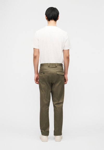 Άνδρας που κοιτάζει προς τα πίσω, φοράει λευκό μπλουζάκι, χακί παντελόνια με πίσω τσέπες και λευκά αθλητικά παπούτσια, σε απλό φωτεινό φόντο.