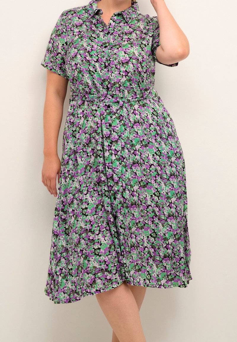 Femme portant une robe fleurie mi-longue avec manches courtes et col, présentant des motifs floraux violets et verts sur un fond clair.