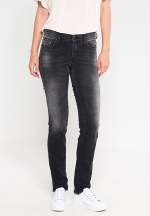 Vrouw die versleten zwarte skinny jeans draagt, witte casual sneakers en een wit T-shirt in haar broek gestopt, staand tegen een effen achtergrond.