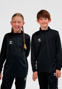 Zwei Kinder tragen identische schwarze Hummel-Sportkleidung und stehen nebeneinander vor einem schlichten weißen Hintergrund.