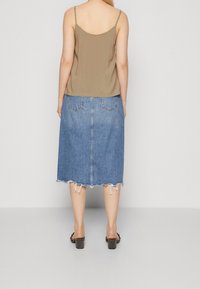 Žena s hnědou topem na ramínka, modrou denimovou sukní po kolena s roztrhaným lemem a černými sandály na podpatku, otočená zády na jednobarevném pozadí.